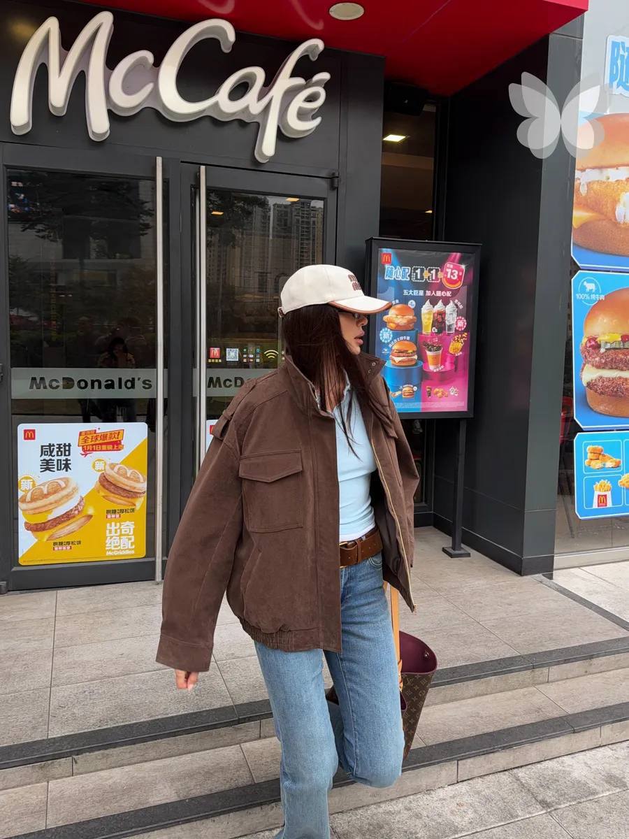 A woman wearing a brown utility jacket, a light blue t-shirt, and light blue jeans walks out of a McDonald's and McCafé restaurant. She is wearing a beige baseball cap and carrying a brown patterned handbag.