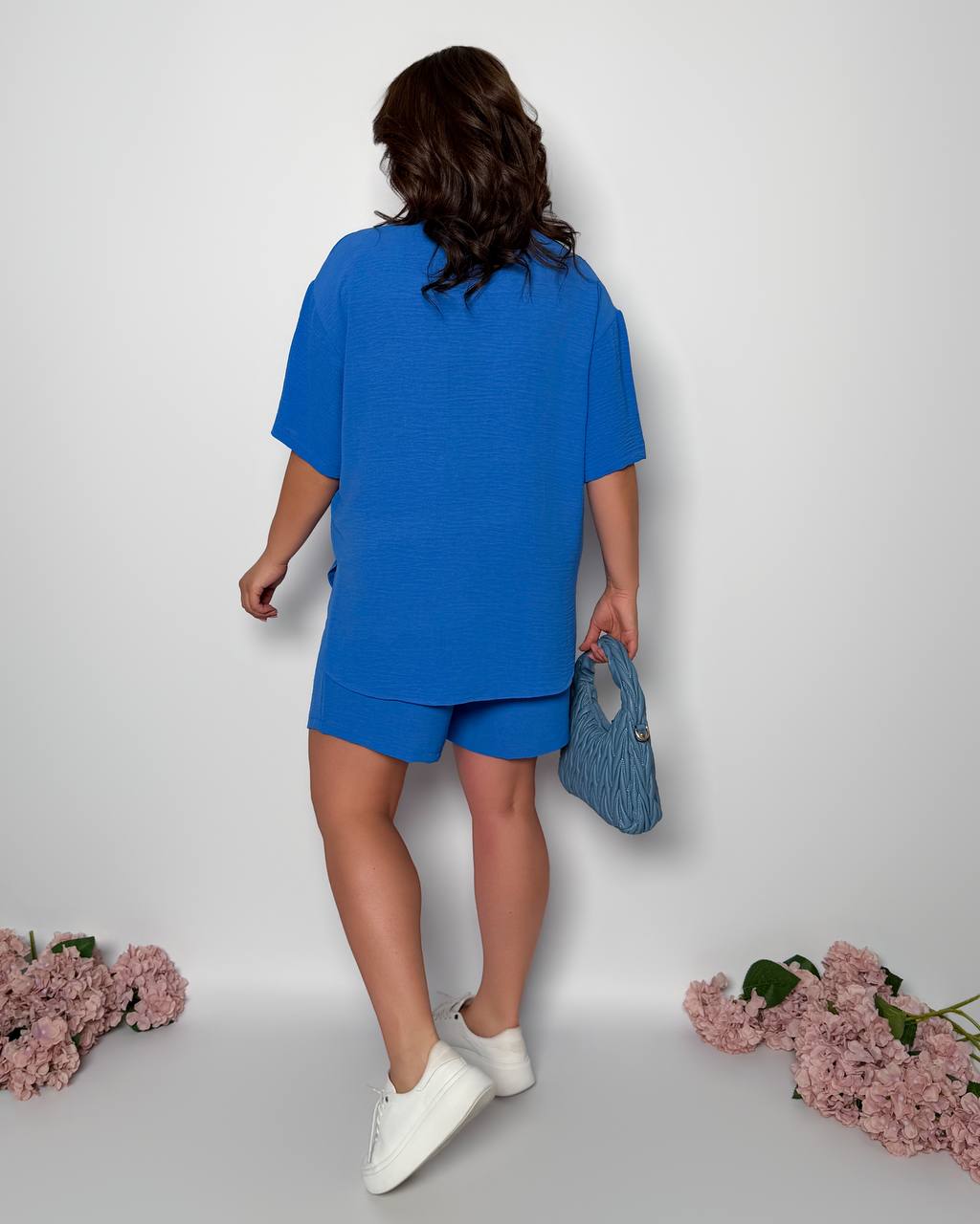 Woman in a blue outfit holding a blue handbag on a light gray background with pink flowers.