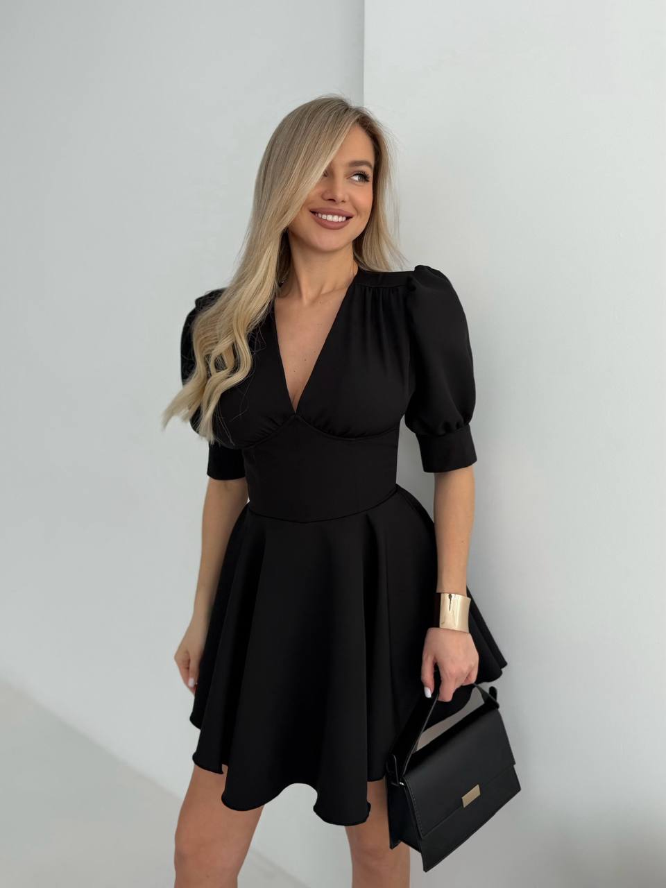 Woman wearing a black dress holding a black handbag against a white wall