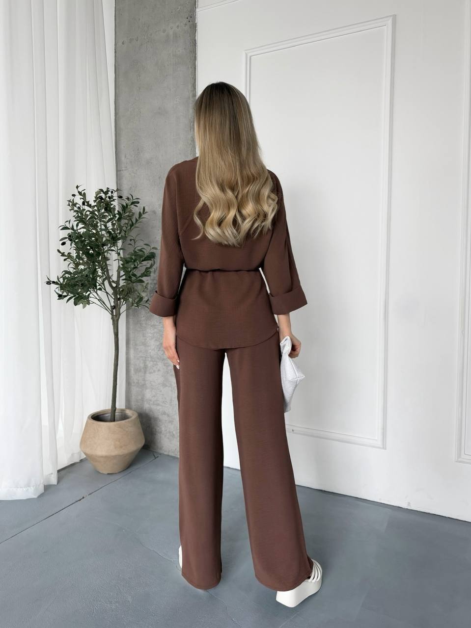 Woman in a brown outfit standing in a room with a plant and white door.