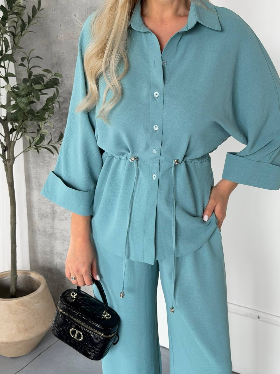 Person wearing a teal outfit with a black handbag indoors.