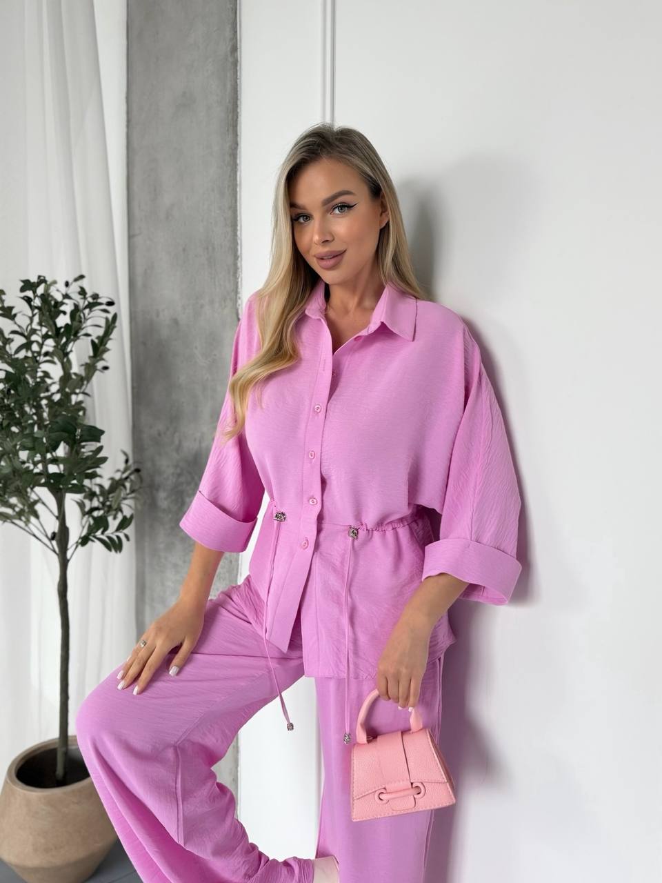 Woman in a pink outfit holding a matching handbag against a white wall.
