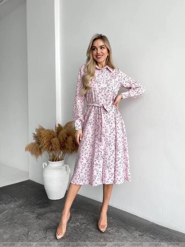 Woman wearing a pink floral dress standing in a minimalistic room with a vase and dried plants.