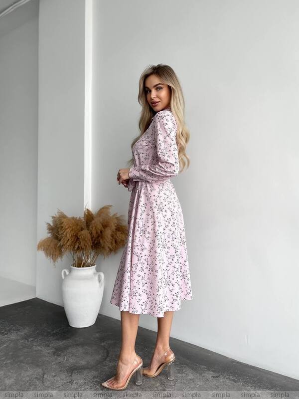 Woman wearing a floral dress standing against a white wall with a vase of dried plants.