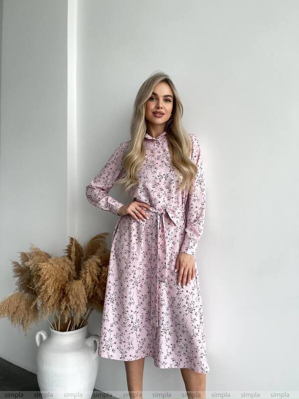 Woman wearing a pink floral dress standing against a white wall with a vase of dried plants.