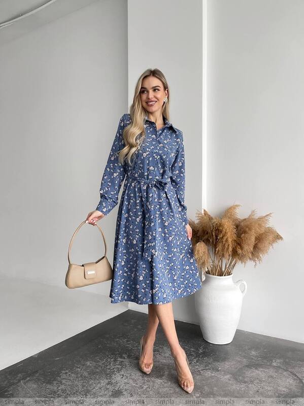 Woman wearing a blue floral dress holding a beige handbag in an indoor setting.