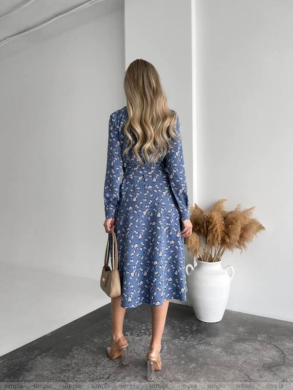 Woman in a blue floral dress standing in a minimalistic room with a vase and dried plants.