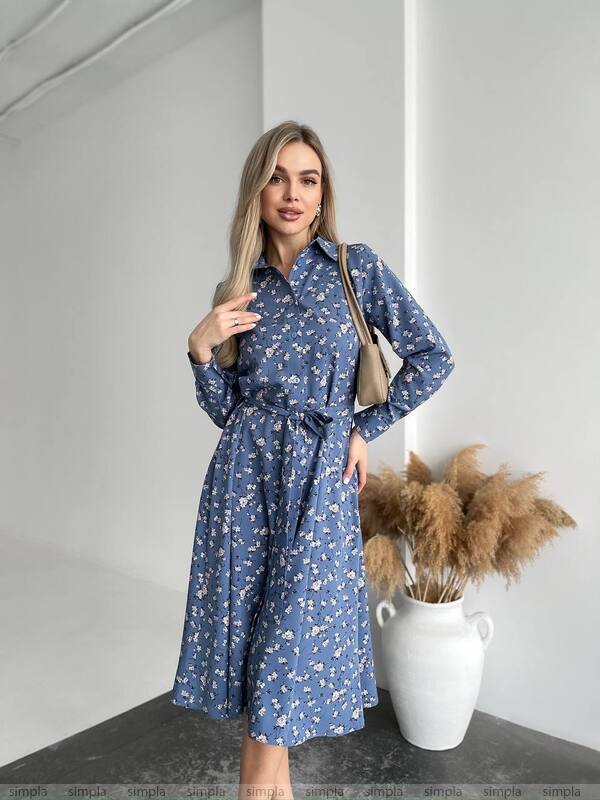 Woman wearing a blue floral dress standing in a minimalistic room with a vase of dried plants.