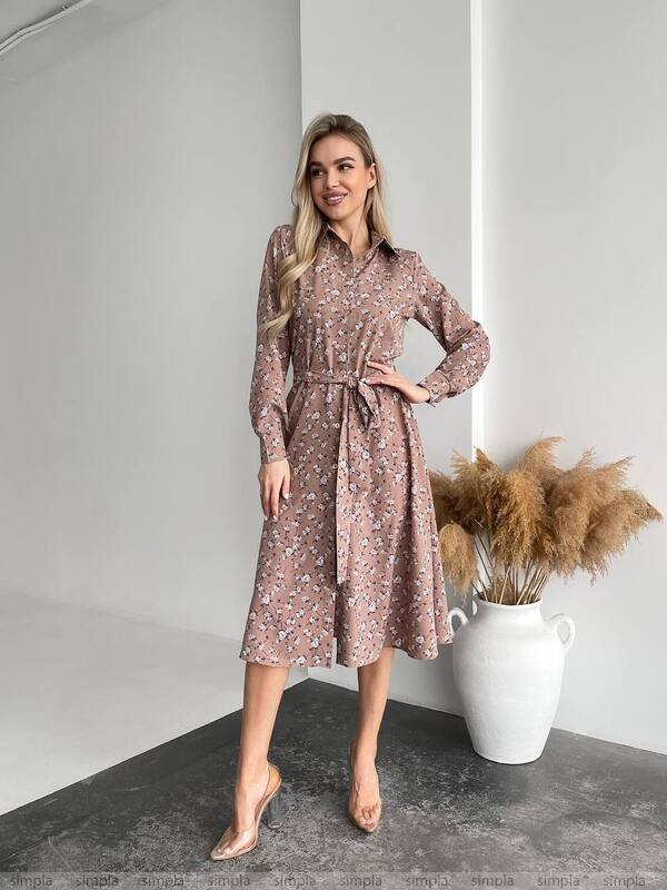 Woman wearing a floral dress standing in a minimalistic room with a vase of dried plants.