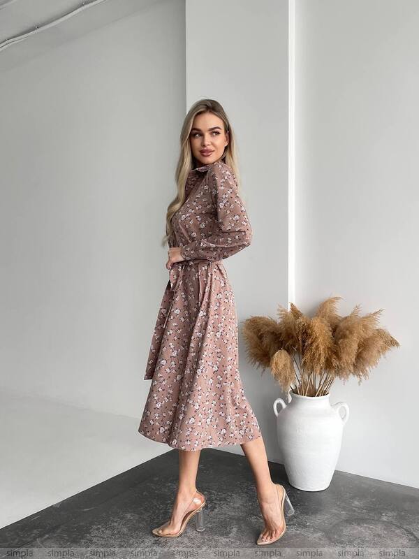 Woman wearing a floral dress standing in a minimalistic room with a vase of dried plants.