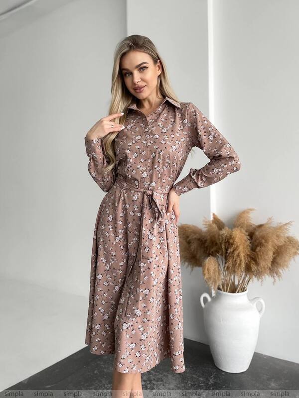 Woman wearing a floral dress standing in a minimalistic room with a vase of dried plants.
