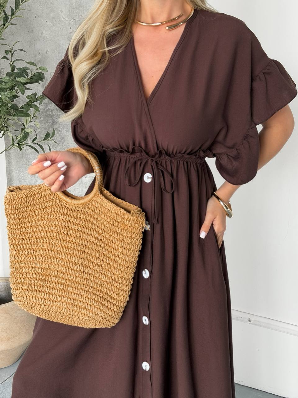 Woman wearing a brown dress holding a straw bag indoors.