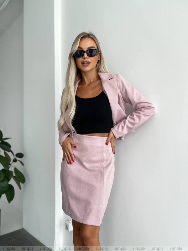 Woman wearing a pink jacket and skirt with a black top, standing against a white wall.