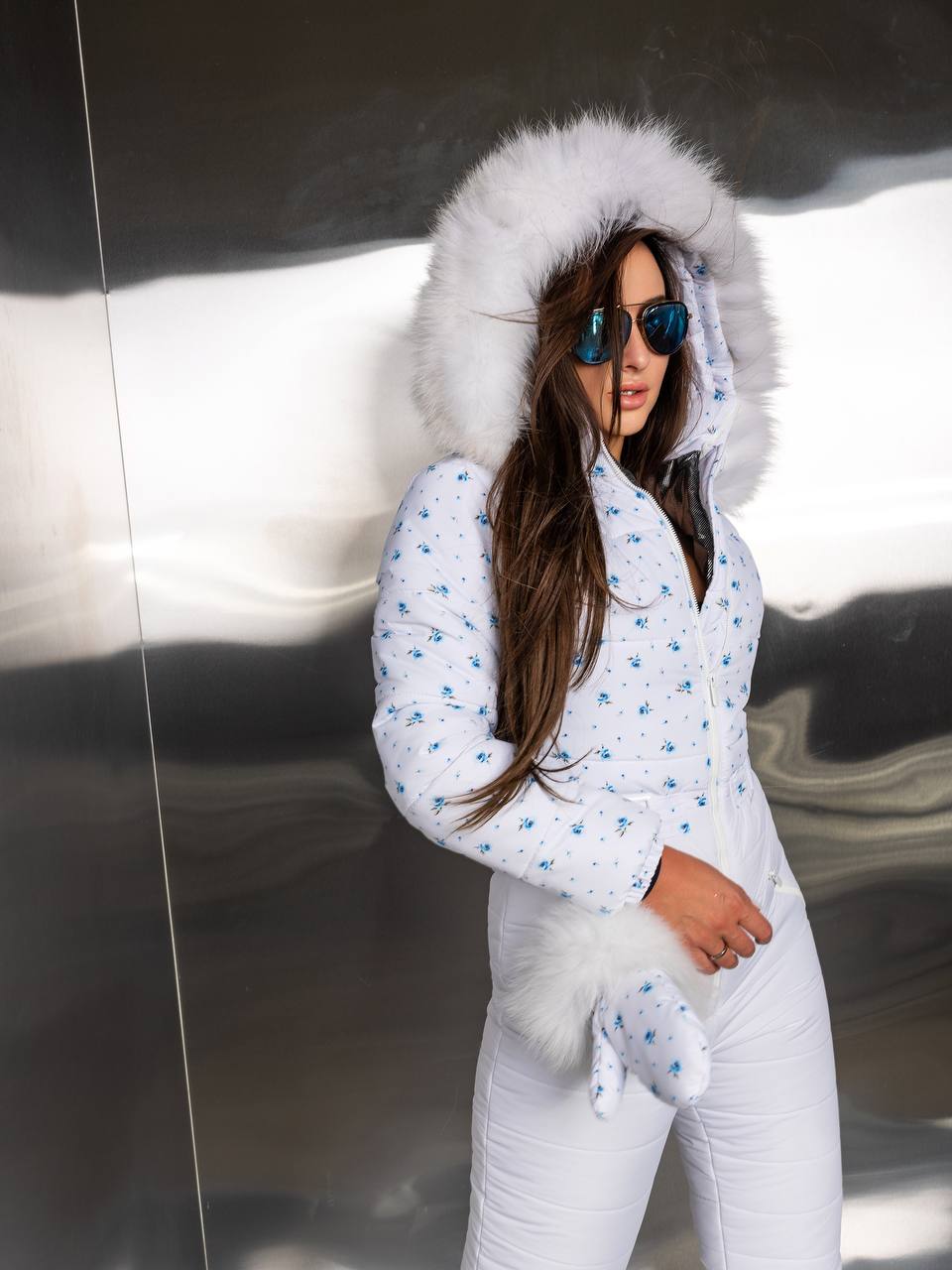Woman wearing a white snowsuit with a fur-lined hood in front of a reflective surface.