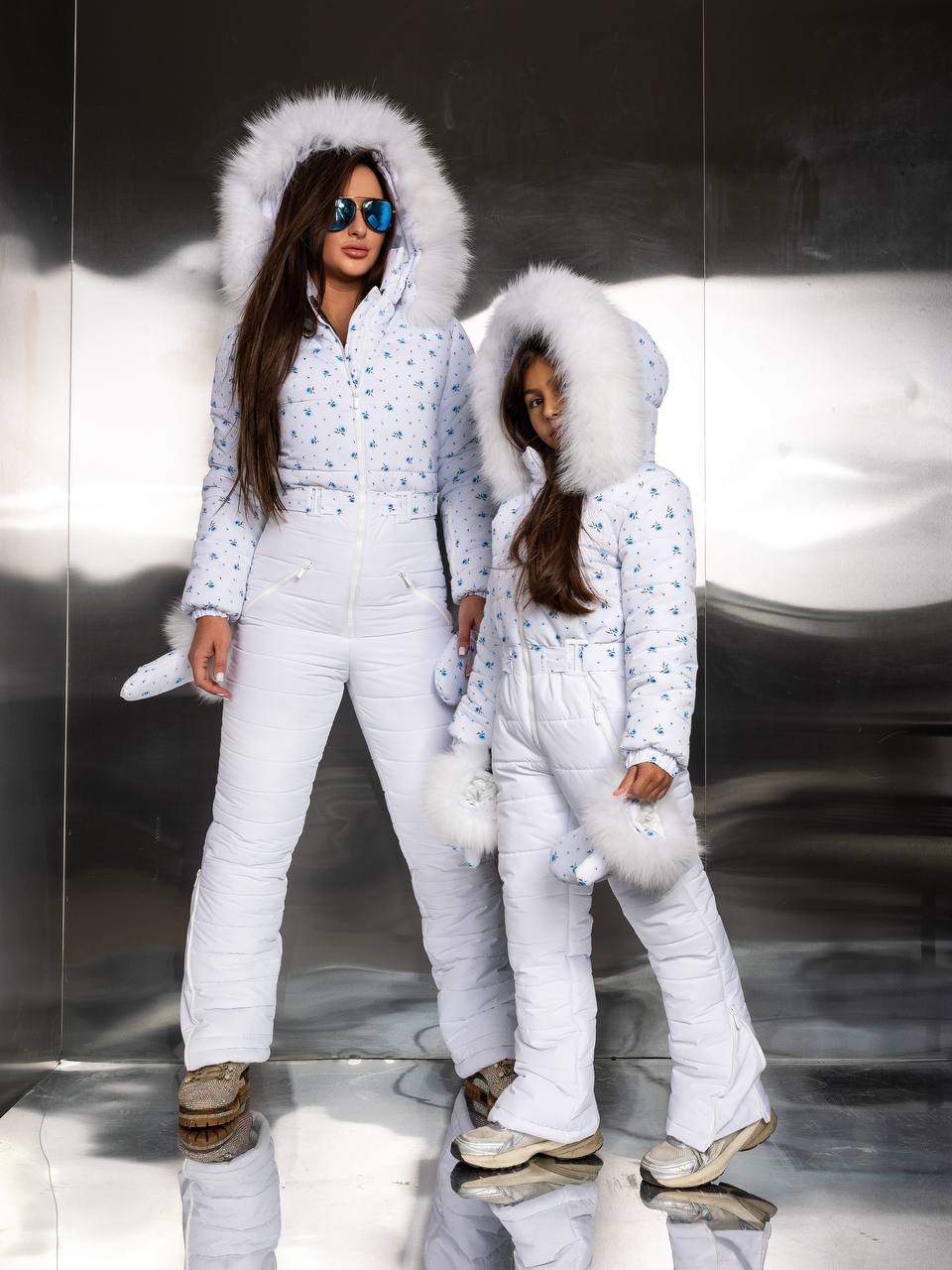 Two people in matching white winter outfits with fluffy hoods on a reflective surface.