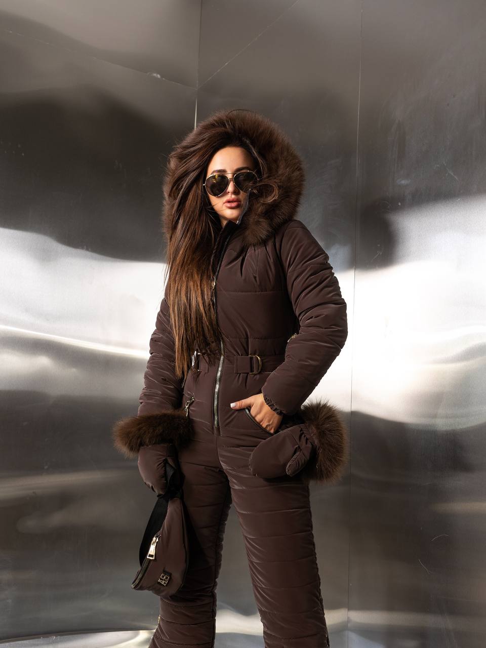 Person wearing a brown fur-lined snowsuit against a reflective metallic wall.