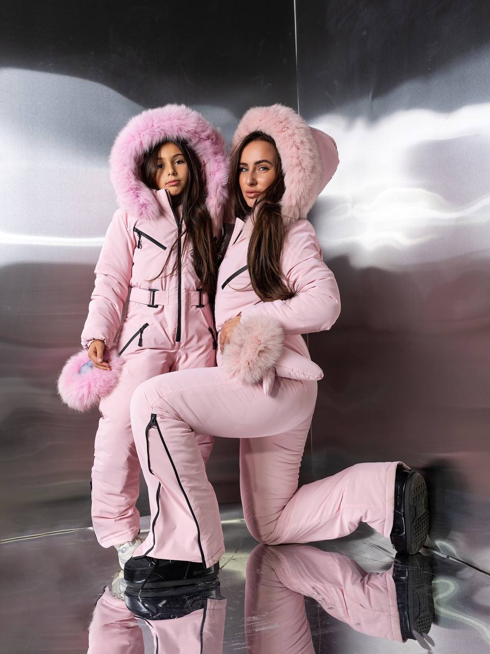 Two women in matching pink winter outfits with fluffy hoods sitting on a reflective surface.