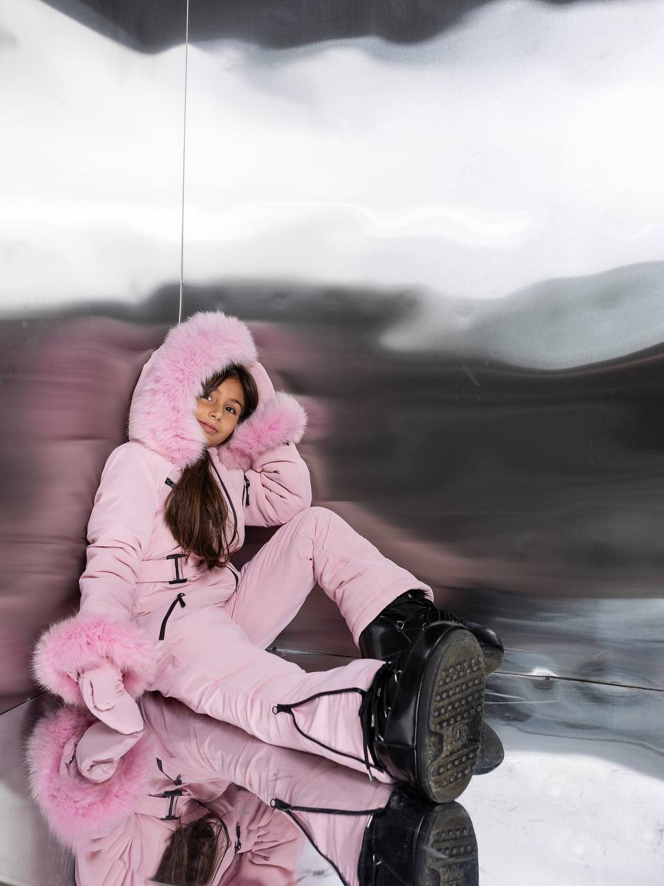 Person wearing a pink snowsuit with a hood sitting on a reflective surface.
