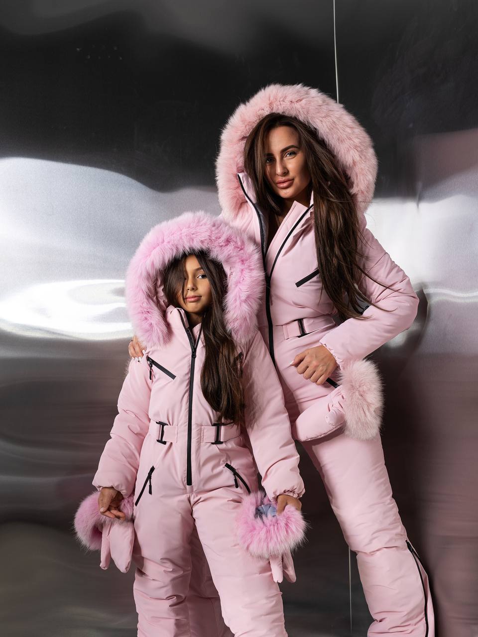 Two people in matching pink outfits with fur hoods against a blurred background