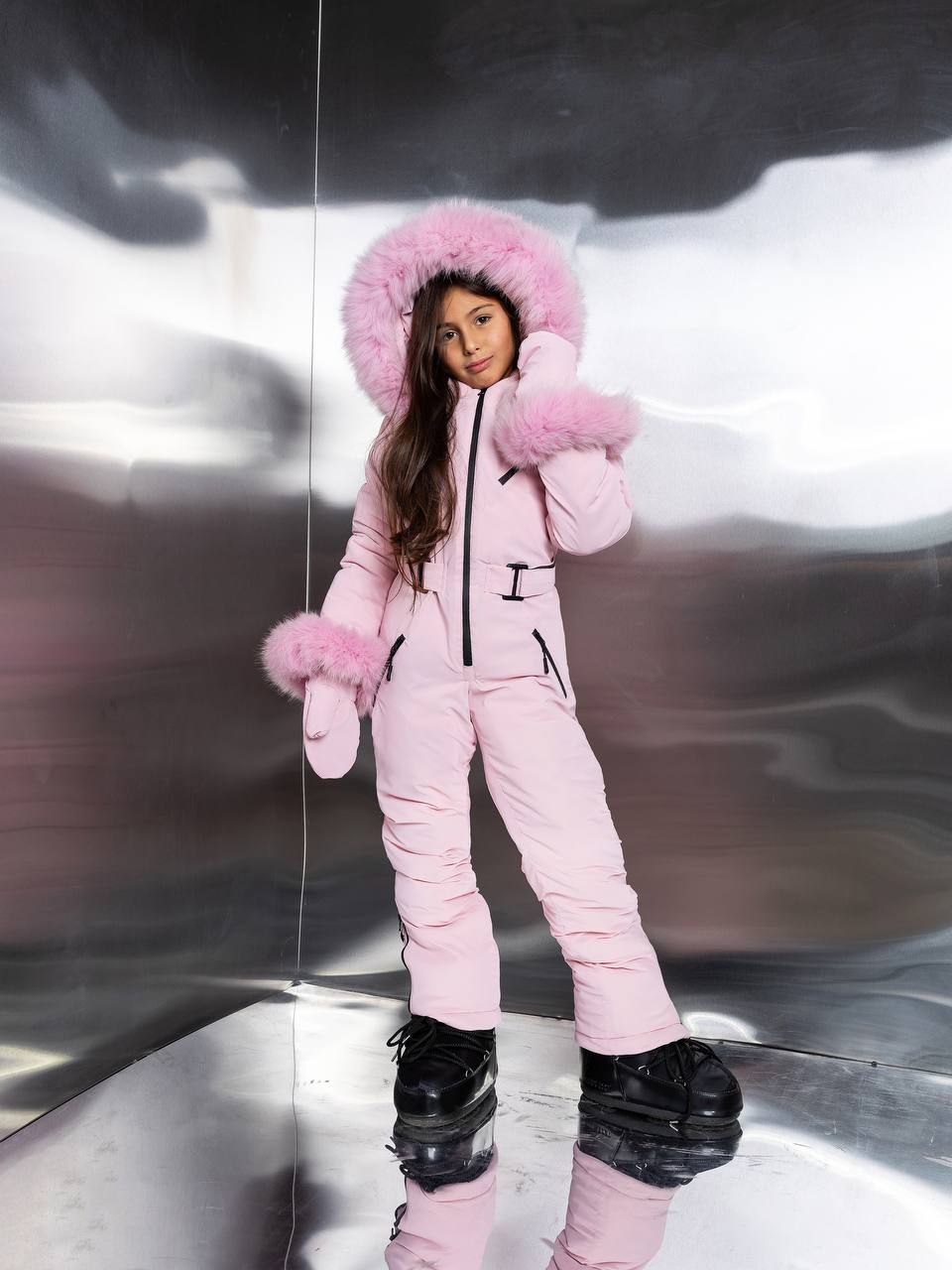 Person wearing a pink snowsuit with fur trim against a reflective surface.