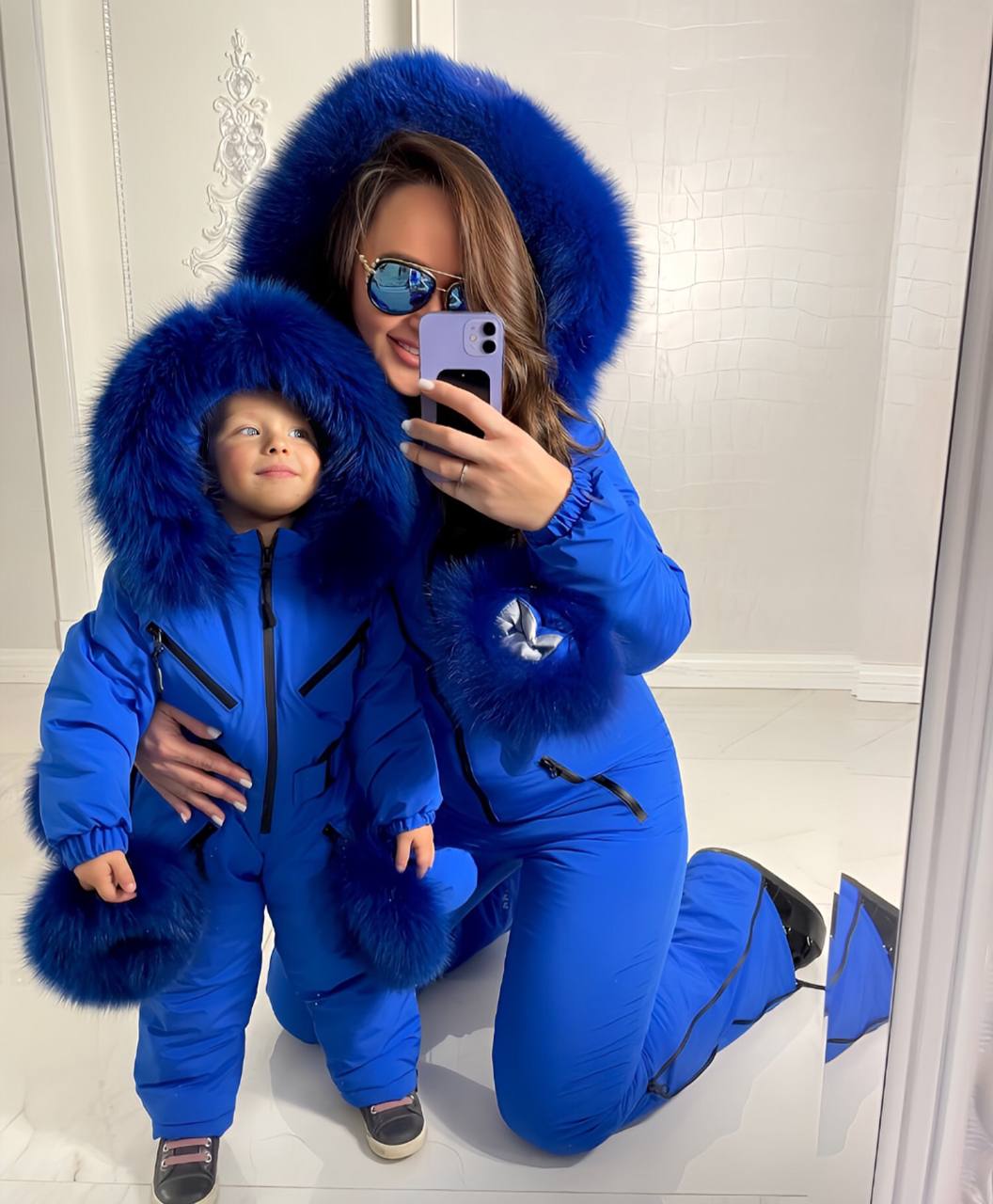 Woman and child in matching blue snowsuits with fur hoods taking a selfie.