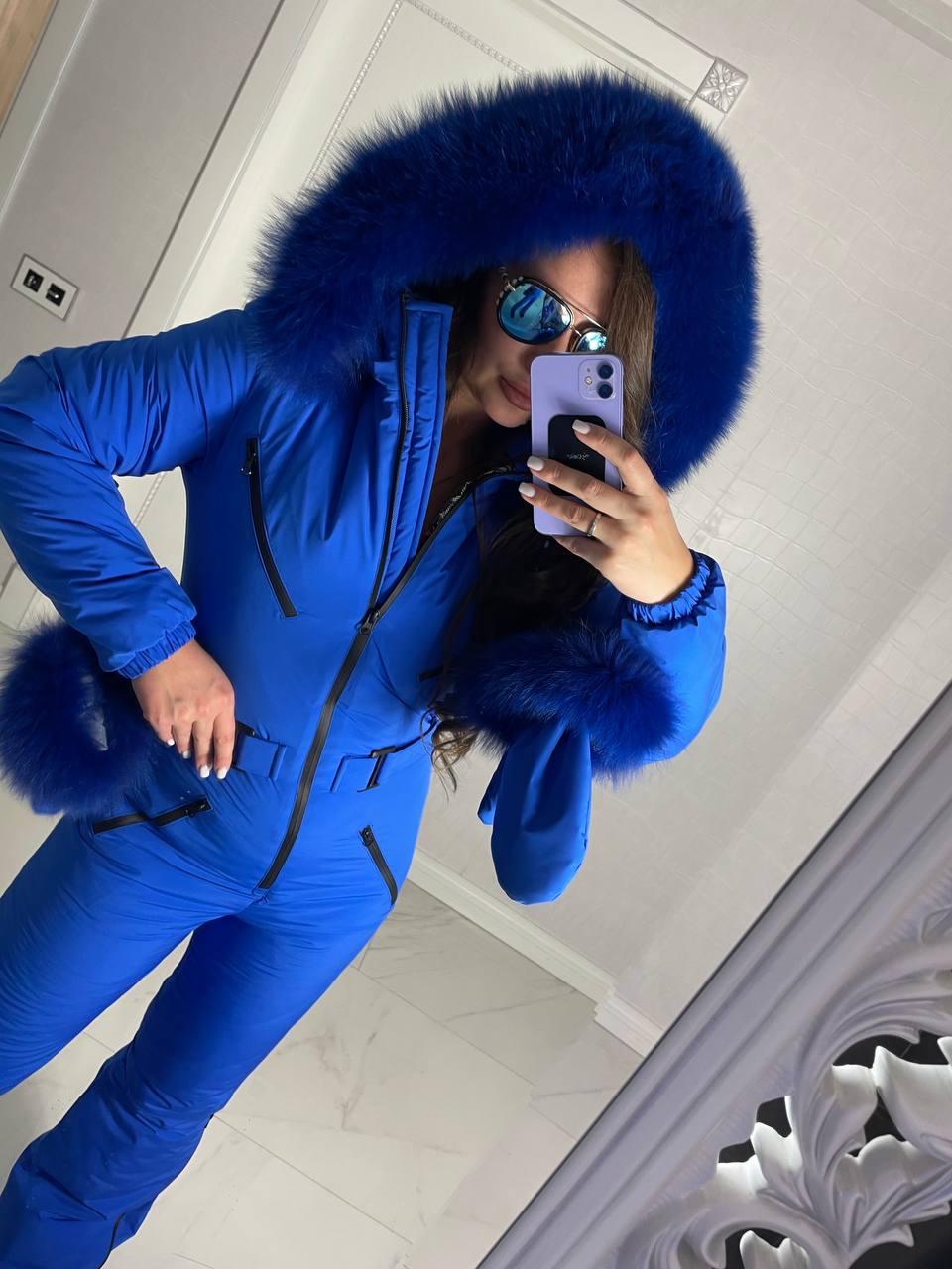 Person wearing a blue snowsuit with a fur-lined hood taking a mirror selfie.