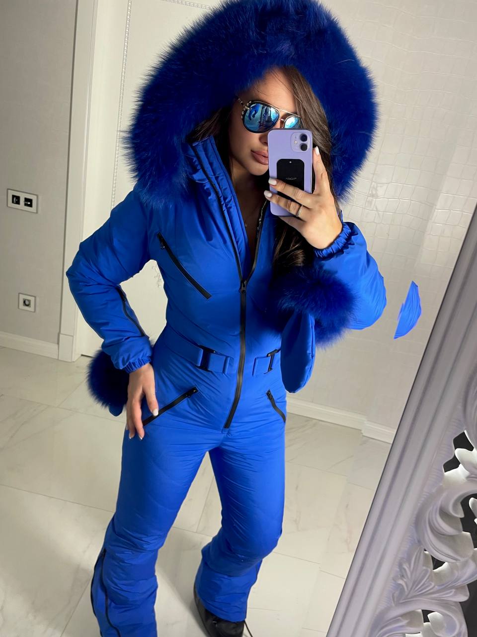 Person wearing a blue snowsuit with a fur-lined hood taking a mirror selfie.