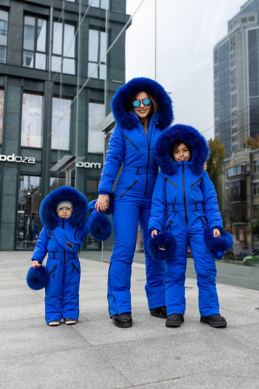 Family of three wearing matching blue snowsuits with fur hoods in an urban setting.