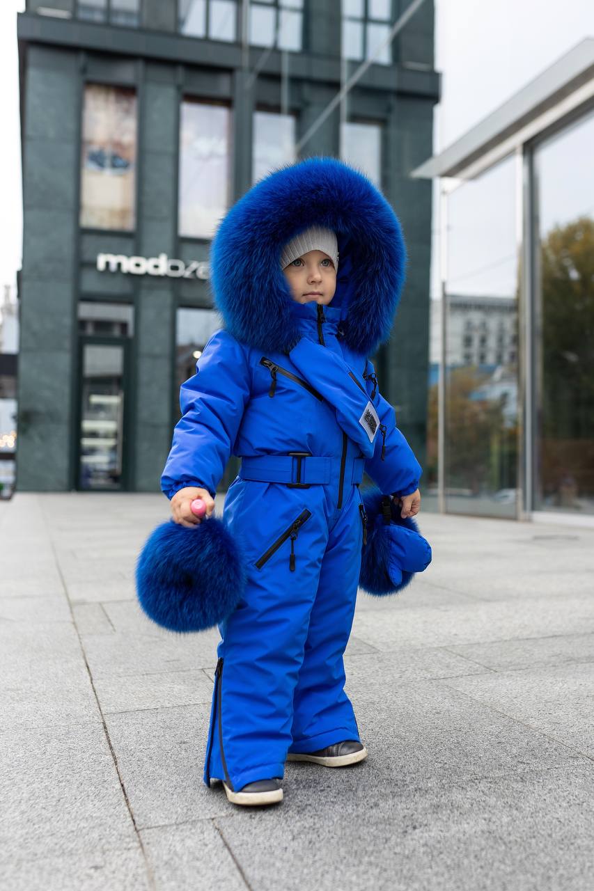 Child in a blue snowsuit with fur hood standing on a sidewalk.