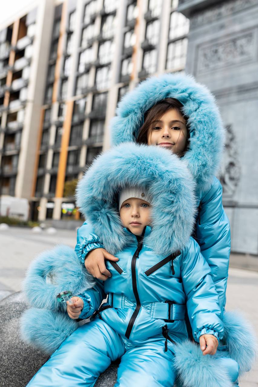 Two children in matching blue fur-lined snowsuits sitting on a street.