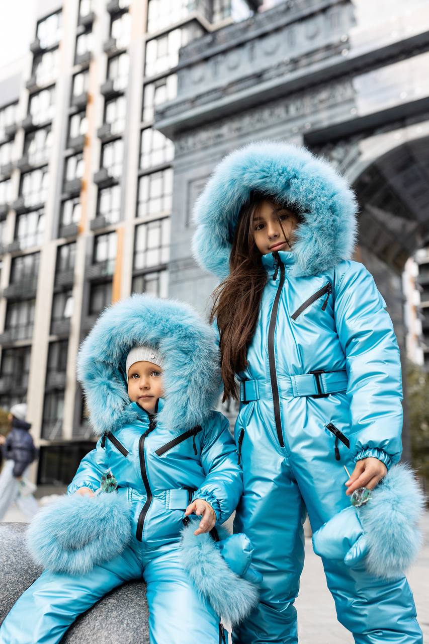 Two children in matching blue snow suits with fur hoods standing in an urban setting.