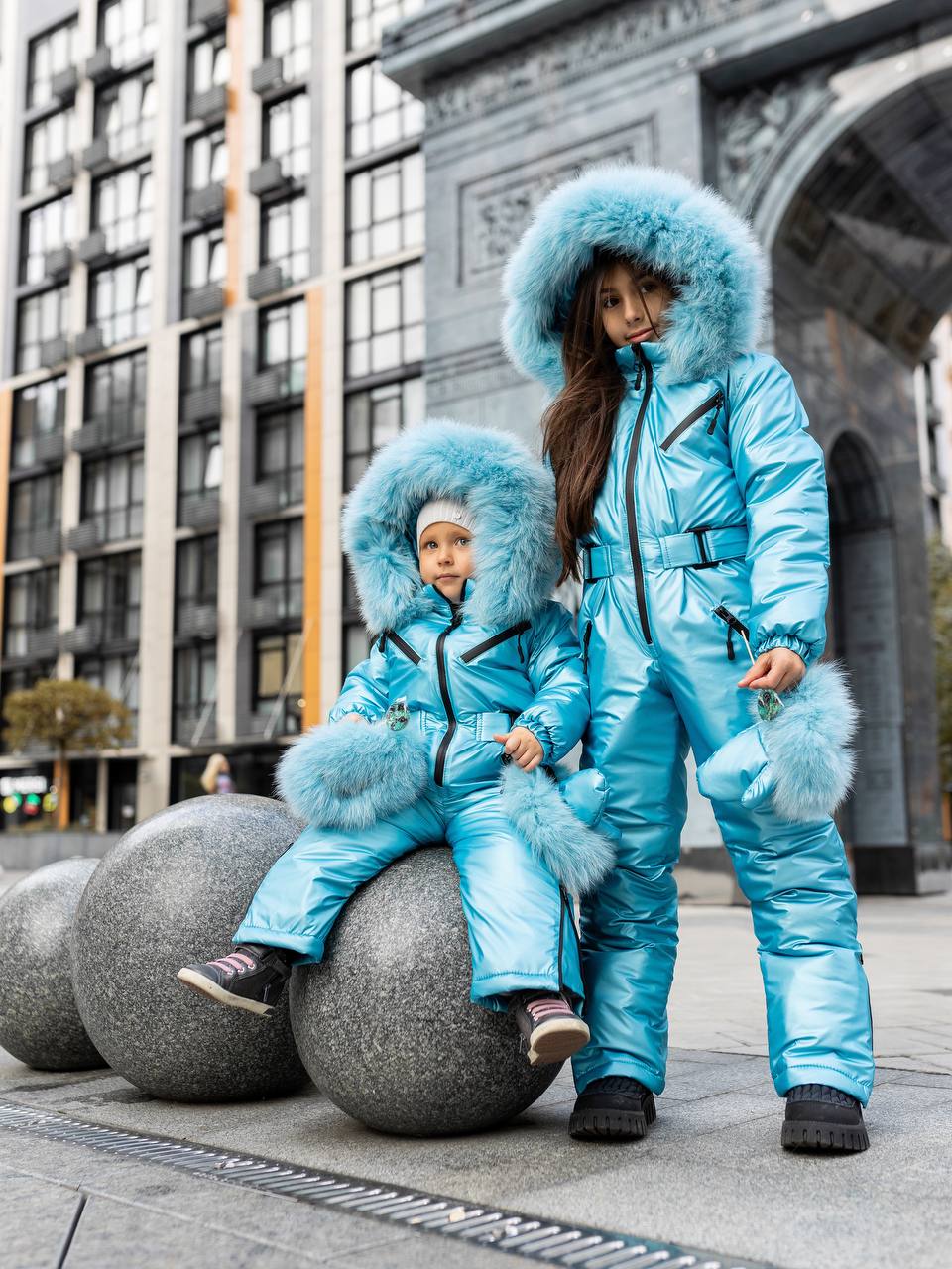 Two children in matching blue snowsuits with fur hoods standing on a city street.