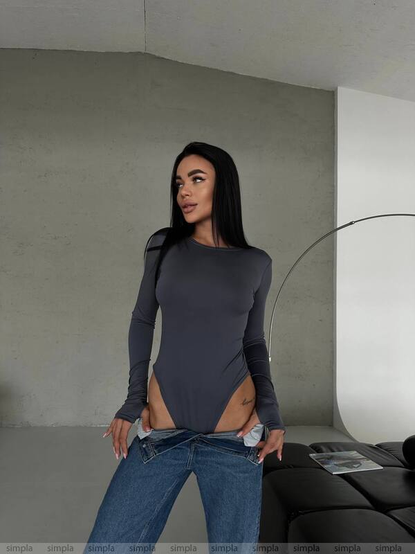 Woman wearing a dark gray long-sleeve top and blue jeans in a minimalistic room.