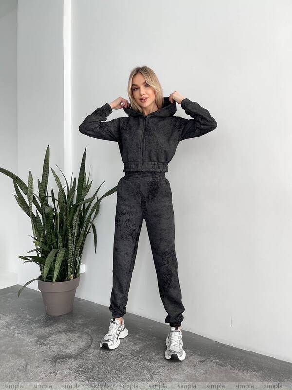 Woman in black tracksuit standing next to a potted plant against a white wall