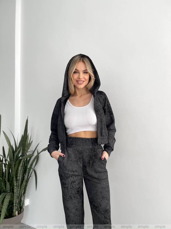 Woman wearing a black hoodie and dark pants standing against a white wall with a plant to the left.