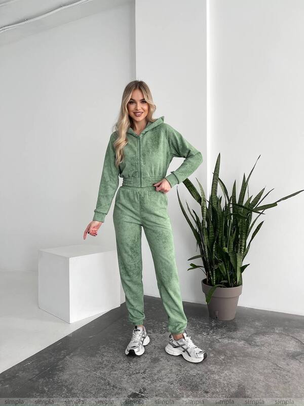 Woman wearing a green tracksuit standing in a minimalistic room with a plant.