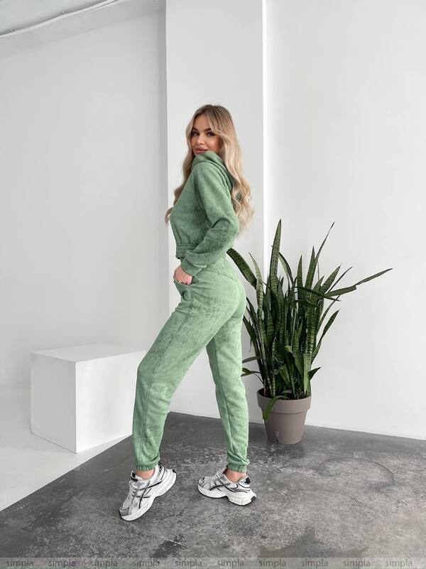 Woman in a green tracksuit standing in a minimalistic room with a plant.