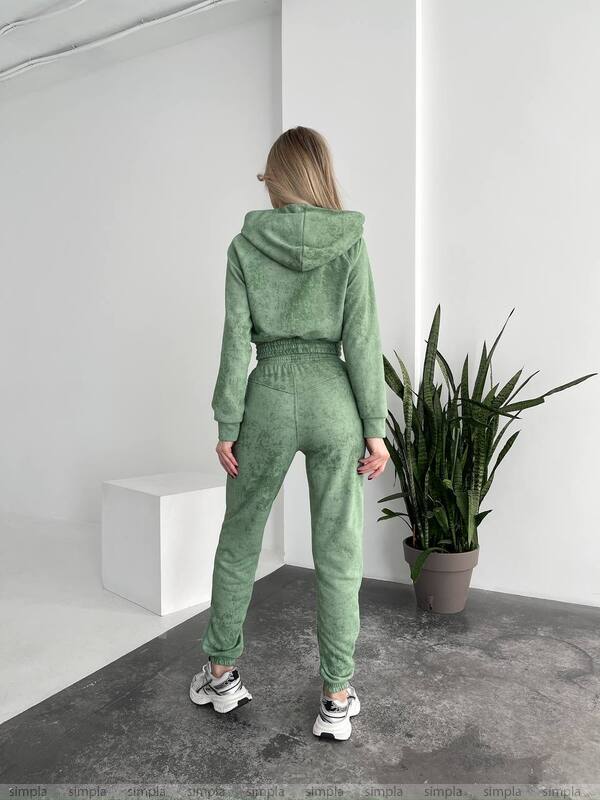 Person wearing a green tracksuit standing in a minimalistic room with a plant.
