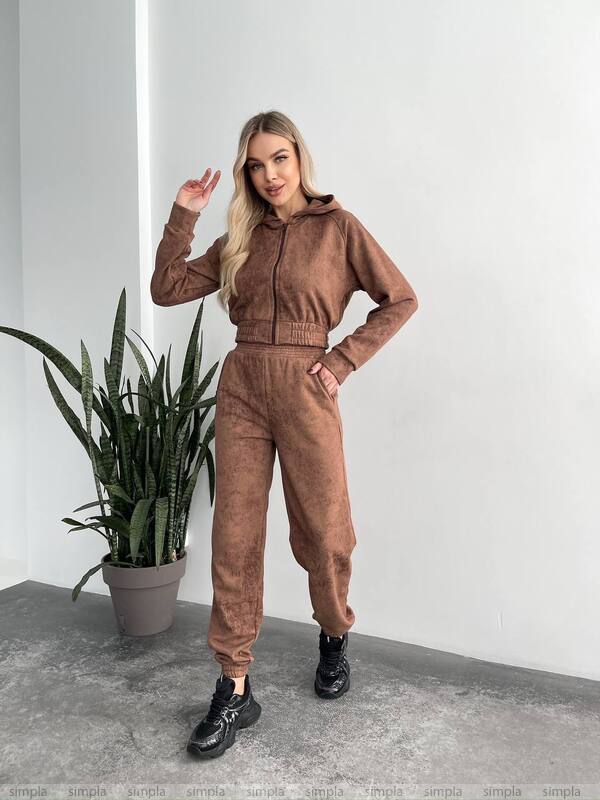 Woman wearing a brown tracksuit standing next to a potted plant against a white wall.