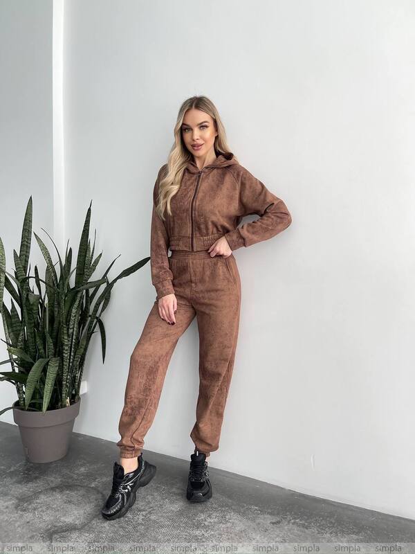 Woman wearing a brown tracksuit standing next to a potted plant against a white wall.