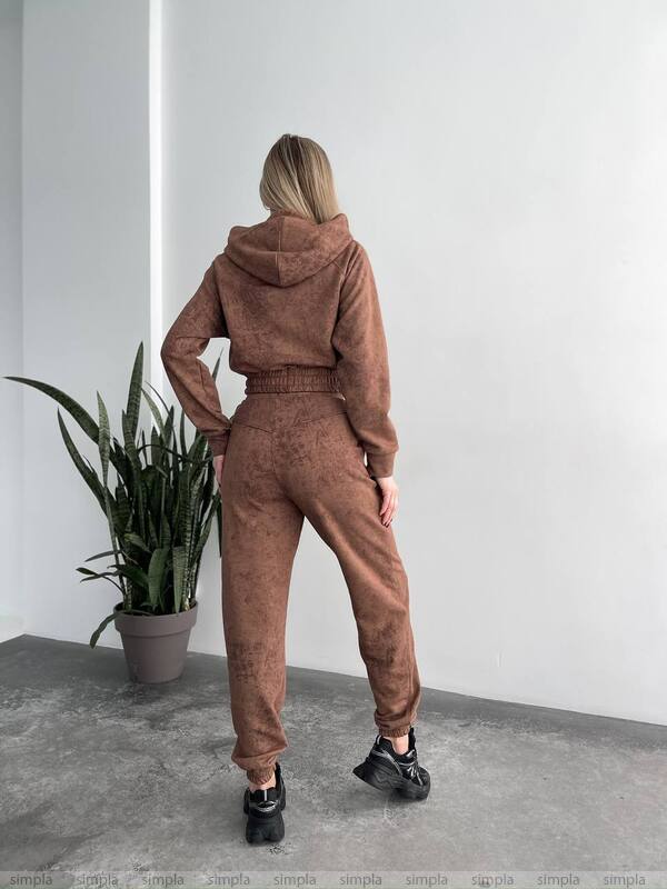 Person wearing a brown tracksuit standing in a minimalistic room with a plant.