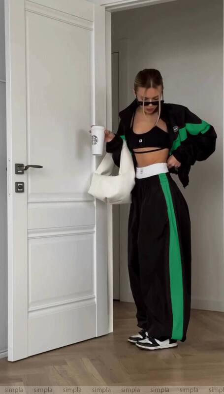 Person in black and green tracksuit with white sports bra and leggings, holding a white bag in a room.
