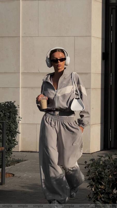 Person in a gray tracksuit with headphones and sunglasses, holding a drink, standing against a light-colored wall.