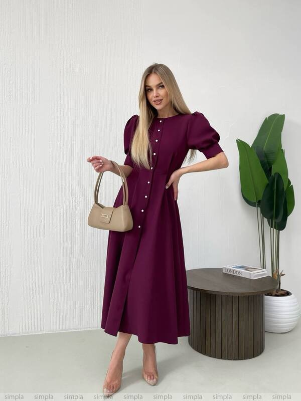 Woman in a burgundy dress holding a beige handbag in a minimalistic room.