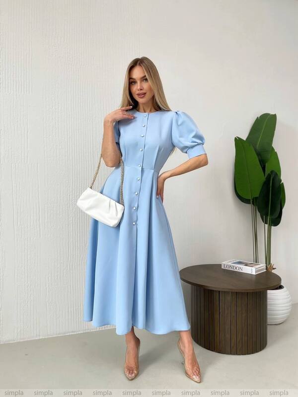 Woman wearing a light blue dress with a white handbag in an indoor setting.
