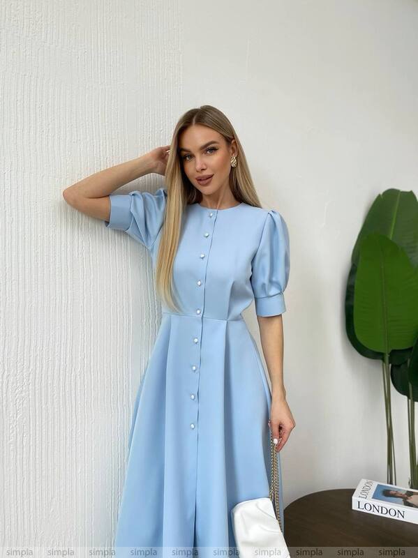 Woman wearing a light blue dress with puff sleeves against a white wall.