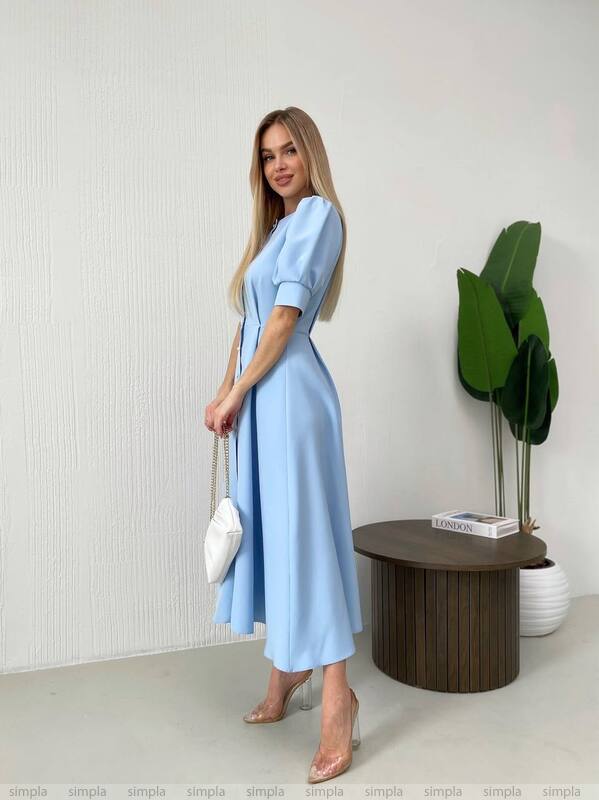 Woman in a light blue dress standing in a minimalistic room with a plant and small table.