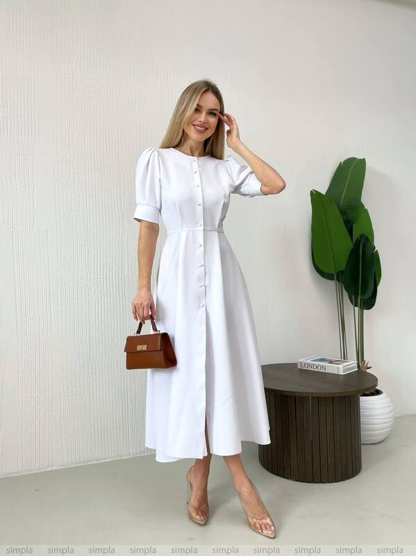 Woman in a white dress standing indoors with a plant and small table in the background