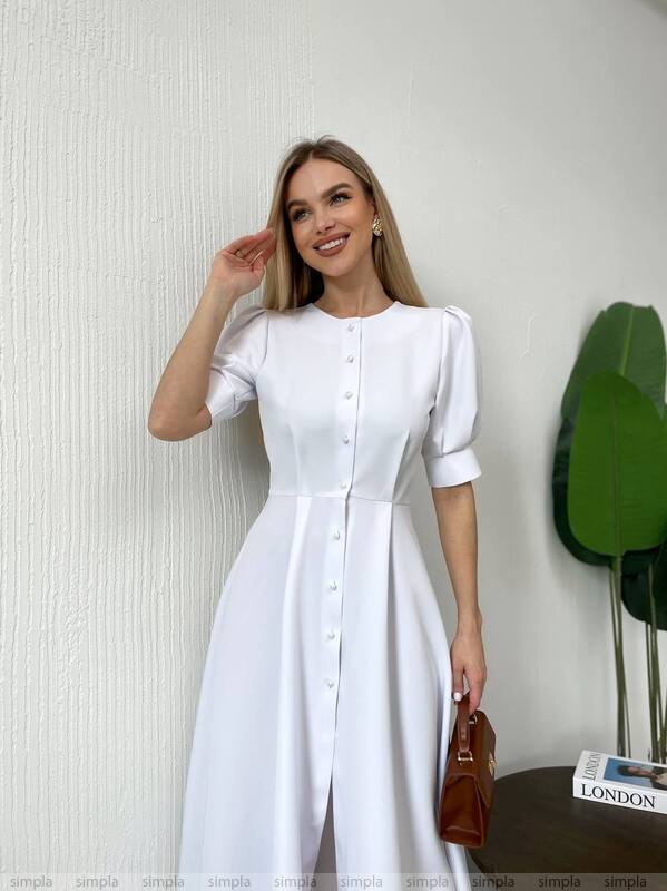 Woman wearing a white dress standing against a plain wall with a plant in the background.
