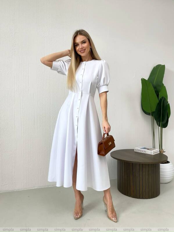 Woman in a white dress standing in a minimalistic room with a plant and small table.
